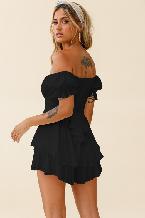 Back 3/4 view of a black off-shoulder summer romper, showcasing the layered ruffles, tie-back closure, and short hemline.
