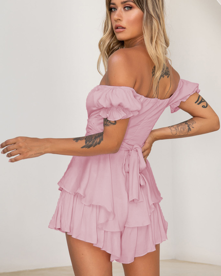 Back 3/4 view of a women's pink off-shoulder tiered ruffle romper, showing the tie-back detail and ruffled shorts.