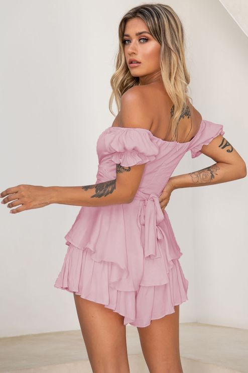 Back 3/4 view of a women's pink off-shoulder tiered ruffle romper, showing the tie-back detail and ruffled shorts.
