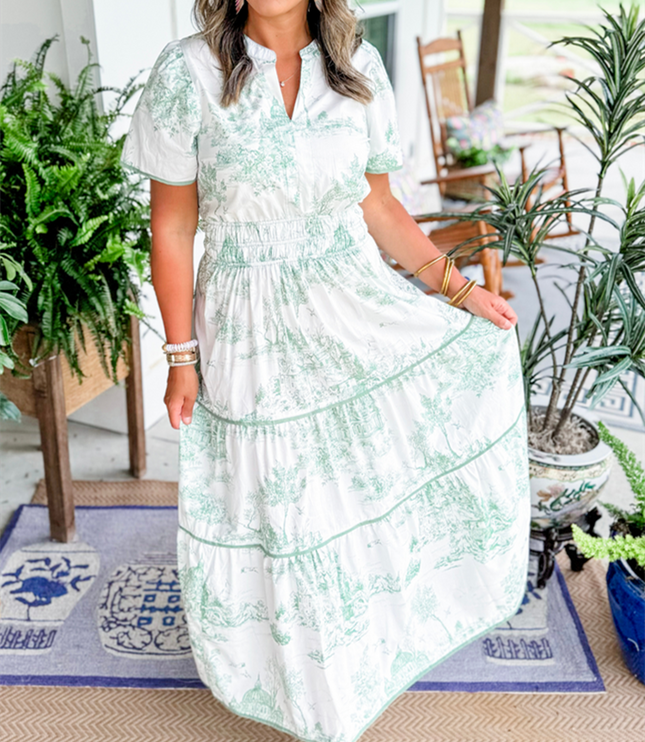 Women's white summer maxi dress with light green scenic print, v-neck, short sleeves, and tiered skirt, posing with hand on skirt.