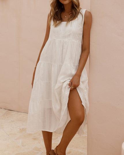 Front view of a woman wearing a white sleeveless tiered midi dress with a square neck and brown flat sandals, leaning against a pink wall.