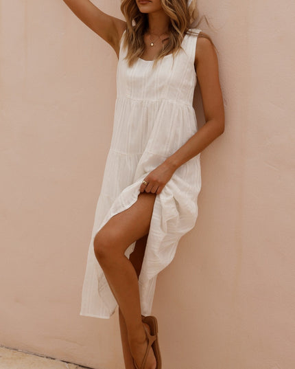 Woman in a white sleeveless tiered midi dress with a square neckline, raising her arm and showing a leg, against a pink wall.