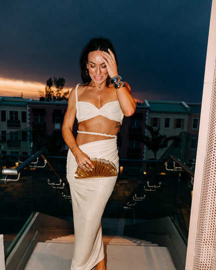 Women's off-white two-piece rope maxi skirt set worn at dusk, featuring the twisted rope details and a gold clutch accessory.