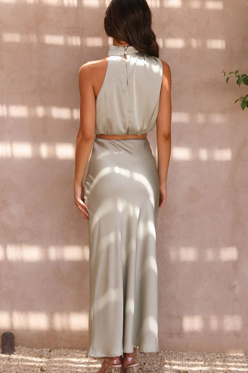 Rear view of a women's light sage green satin two-piece set featuring an asymmetrical high neck crop top and long skirt.