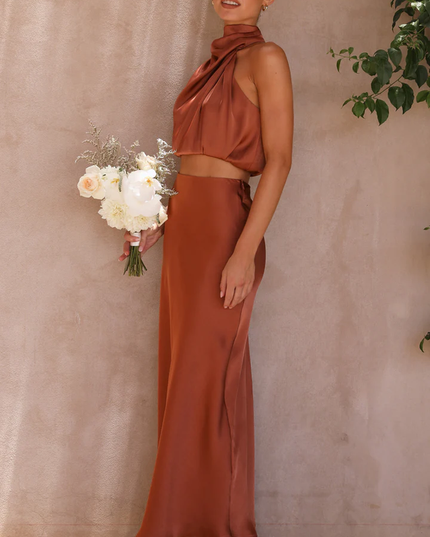 Side view of a women's rust orange satin asymmetrical high neck crop top and maxi skirt set, holding a white bouquet.
