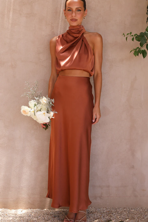 Front view of a women's rust orange satin high neck crop top and maxi skirt set, holding a white floral bouquet.