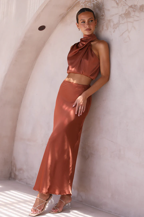 Women's rust orange satin asymmetrical high neck crop top and maxi skirt set, leaning against a wall, showcasing the flowy fabric.
