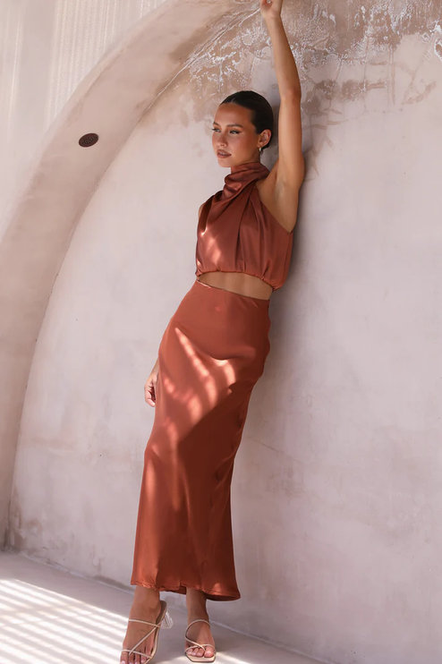 Full length view of a women's rust orange satin high neck crop top and maxi skirt set, with arm raised, suitable for wedding guests.