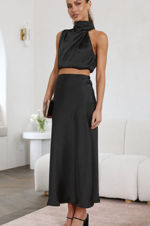 Side view of a woman in a black satin asymmetrical high neck crop top and matching maxi skirt, with black block heels.