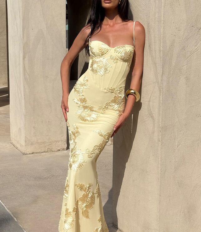 Full-length side view of a woman in a light yellow floral corset maxi dress with gold floral embroidery, posing outdoors.