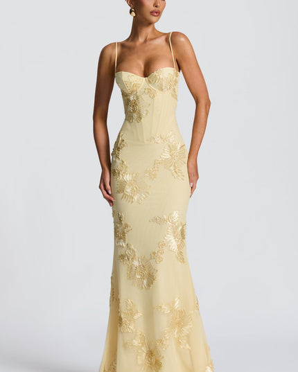 Frontal view of a woman standing in a light yellow floral corset maxi dress, featuring gold floral embroidery and a fitted silhouette.