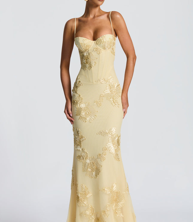 Frontal view of a woman standing in a light yellow floral corset maxi dress, featuring gold floral embroidery and a fitted silhouette.