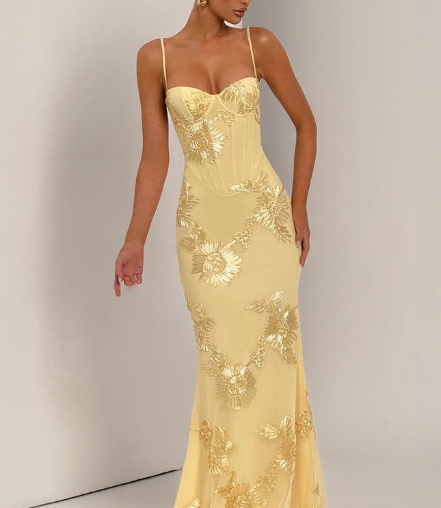 Full-length view of a woman in a light yellow floral corset maxi dress with gold embroidery, posing with one arm outstretched.