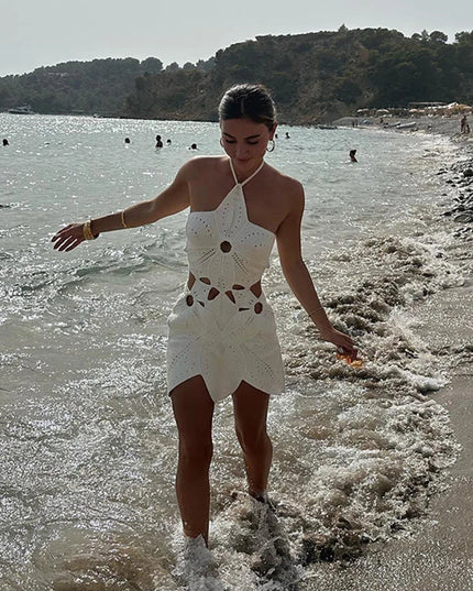 Women's white crochet halter neck mini dress with cut-outs, walking along a beach shoreline with waves.