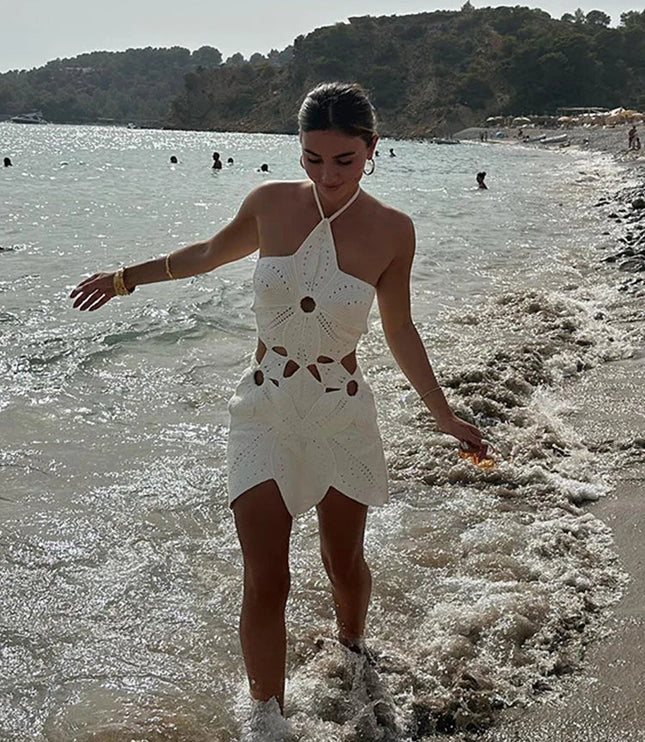 Women's white crochet halter neck mini dress with cut-outs, walking along a beach shoreline with waves.