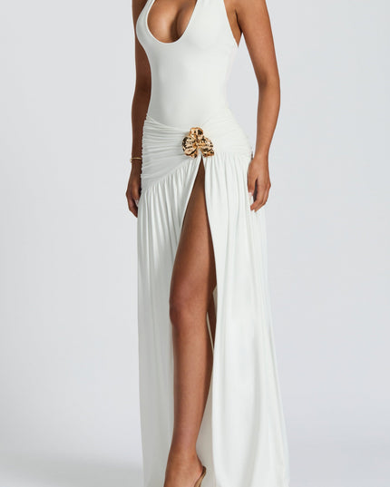 Side profile of a women's white high waist ruched split maxi skirt with a gold floral buckle, worn with a white halter neck top and nude heels.