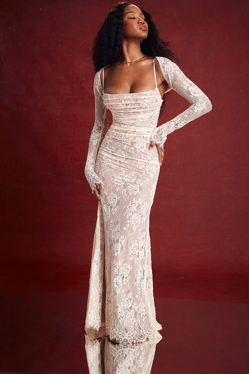 White lace strapless maxi dress with long sleeve bolero, ruched bodice, and floral lace overlay, against a red backdrop.