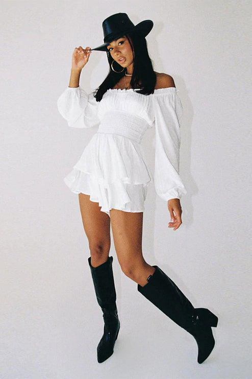 Full view of white off-shoulder mini dress with long sleeves and ruffled hem, accessorized with a black hat and tall black cowboy boots.