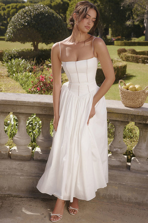 Outdoor shot of women's white sleeveless midi dress, featuring a square neckline and full pleated skirt, with white heels.